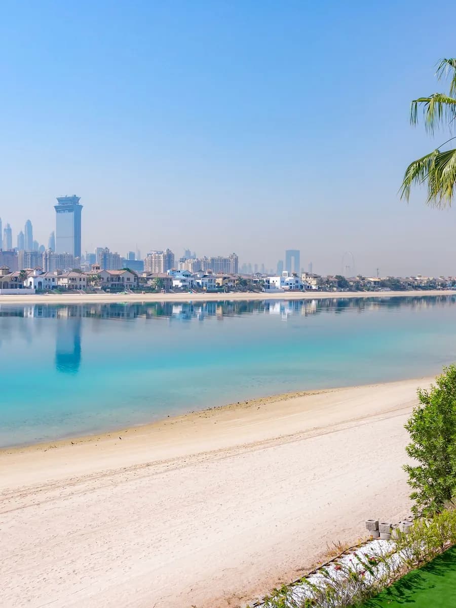 Palm Jumeirah plajında turkuaz deniz, açık renk kum, palmiyeler ve uzakta Dubai silueti
